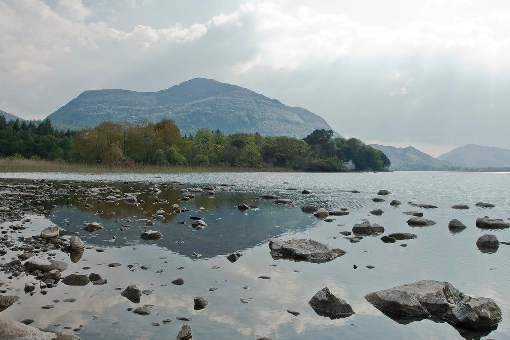 Shallow waters and rocky shoreline of Muckross Lake with Torc Mountain rising beyond the wooded banks in Killarney National Park.