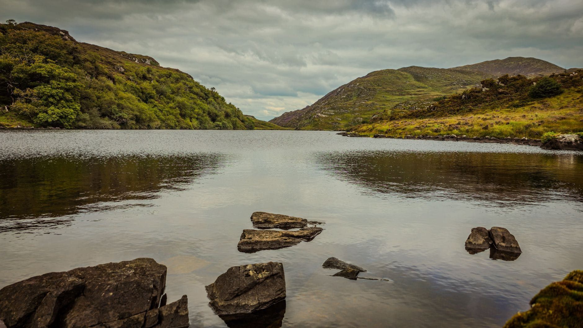 Ruhiges Wasser eines Sees, das sanfte grüne Hügel und bewölkten Himmel im Killarney-Nationalpark widerspiegelt.
