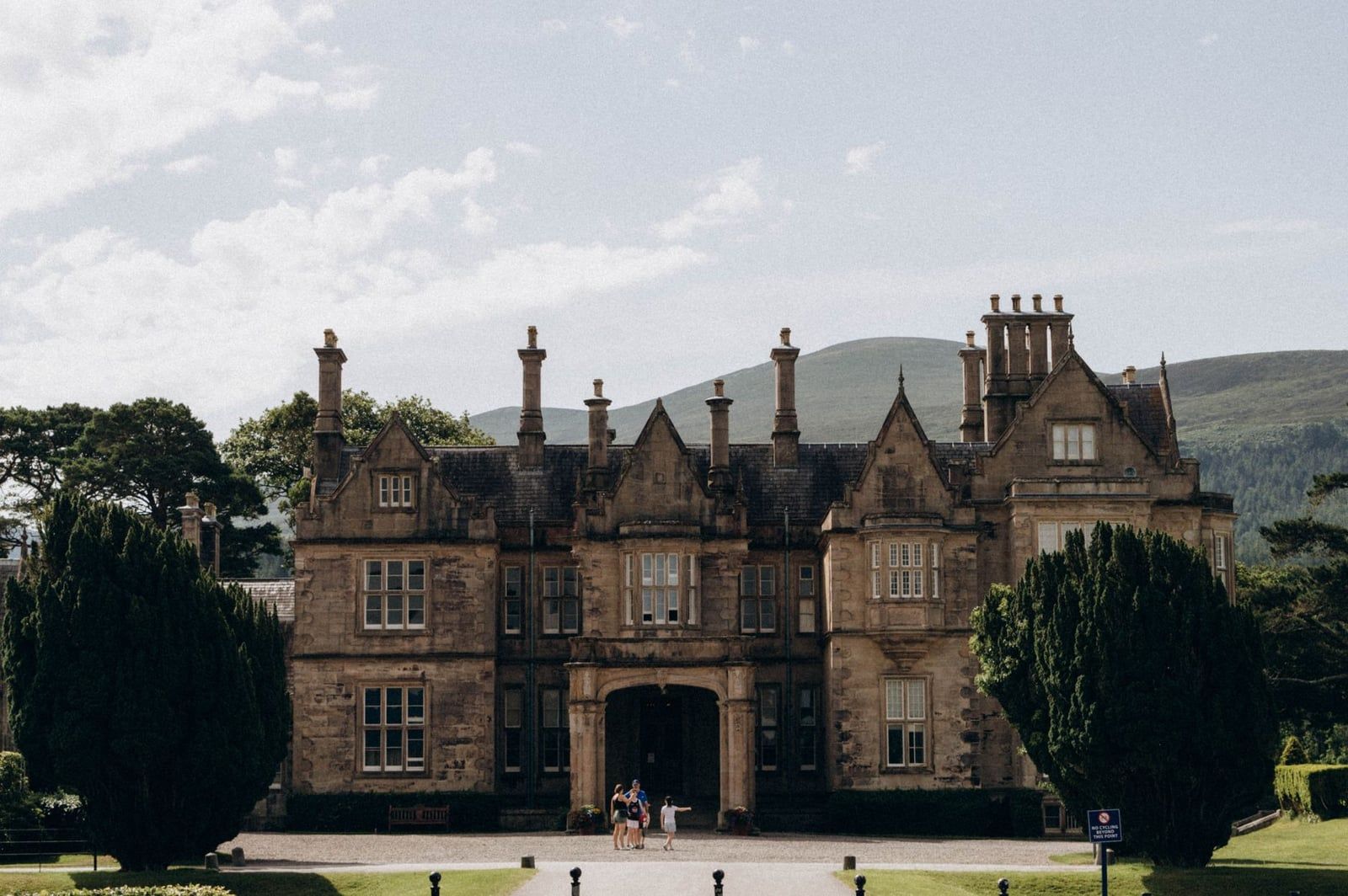 Visitors stand before the Victorian stone facade of Muckross House against a backdrop of mountains in Killarney National Park.